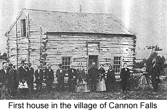 Black and white photo of a rough frame cabin, before which stand several men and women in mid-19th. century clothing--the first house in the village of Cannon Falls