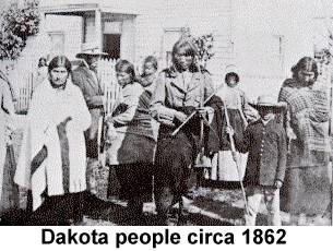 Black and white photo of several Dakota Sioux men, women and children grouped before some frame buildings in 1862