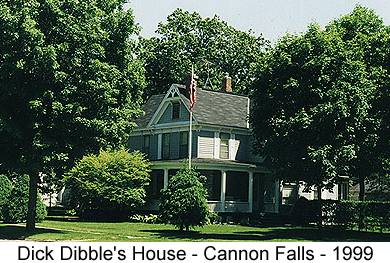 IMAGE/PHOTO: Dick Dibble's House - Cannon Falls - 1999: Color photo of a blue two-story late-Victorian frame house with a high 3rd floor attic and wide wrap-around front porch. The house is surrounded by large trees and shrubs and a white flagpole in the front yard displays the Stars and Stripes hanging loosely in the still air.