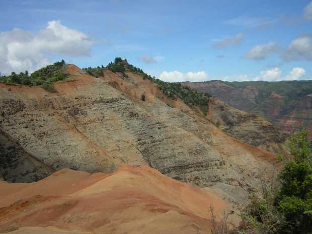 A rough blanket of red sandy rock leads toward a row of grey=banded hills splashed with rusty orange and topped with vegetation. A rough blanket of red sandy rock leads toward a row of grey=banded hills splashed with rusty orange and topped with vegetation.