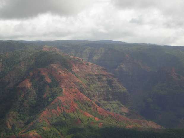 Under a heavy leaden gray sky, a rumpled blanket of red peaks tufted with rough green vegetation stands before distant dark brown and grey canyon walls. Under a heavy leaden gray sky, a rumpled blanket of red peaks tufted with rough green vegetation stands before distant dark brown and grey canyon walls.