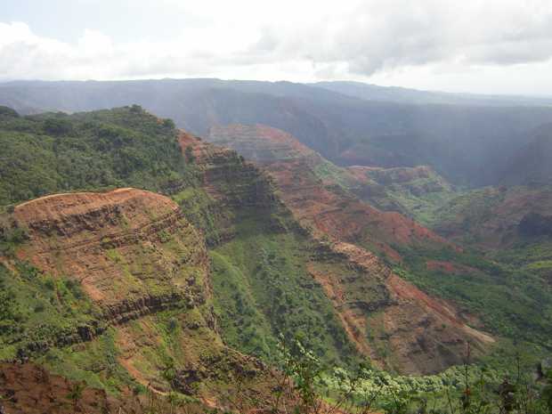 A sheer cliff face, some of its horizontal layers of red rock exposed, the rest covered by vegetation, runs outward from the left and drops off; beyond it rough red and green hills fade into misty blue. A sheer cliff face, some of its horizontal layers of red rock exposed, the rest covered by vegetation, runs outward from the left and drops off; beyond it rough red and green hills fade into misty blue.