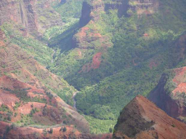 Dimly seen at the bottom of a narrow, steep valley, a brown stream zigzags from top left, through wooded banks and over rapids, before being hidden by red, shrub-dotted hills. Dimly seen at the bottom of a narrow, steep valley, a brown stream zigzags from top left, through wooded banks and over rapids, before being hidden by red, shrub-dotted hills.