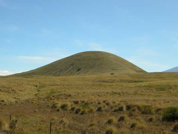 Against a nearly cloudless blue sky, an almost perfectly rounded, greenish tan hill rises suddenly from a nearly flat dry prairie of tufted grass. Against a nearly cloudless blue sky, an almost perfectly rounded, greenish tan hill rises suddenly from a nearly flat dry prairie of tufted grass.