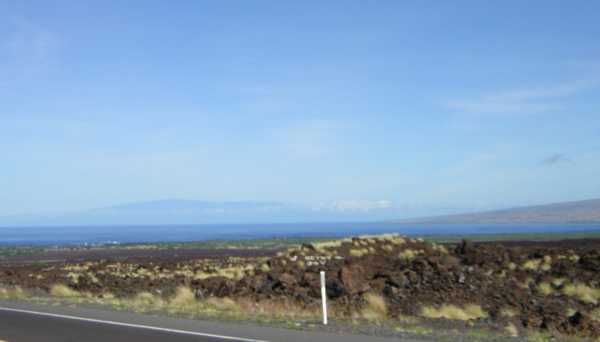 From a roadside, jagged reddish-brown rocky chunks interspersed with yellow-tan tufts of dry grass stretch away to the distant sea, beyond which a shadowy blue mountain rises. From a roadside, jagged reddish-brown rocky chunks interspersed with yellow-tan tufts of dry grass stretch away to the distant sea, beyond which a shadowy blue mountain rises.