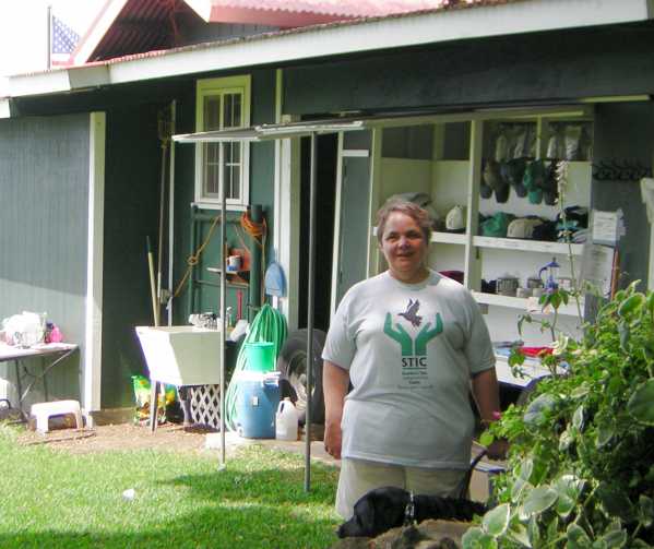 Maria, in STIC t-shirt and tan shorts, stands smiling, with Bella, in front of an outdoor t-shirt and cap display against the back wall of the Greenwell Farms store. Maria, in STIC t-shirt and tan shorts, stands smiling, with Bella, in front of an outdoor t-shirt and cap display against the back wall of the Greenwell Farms store.