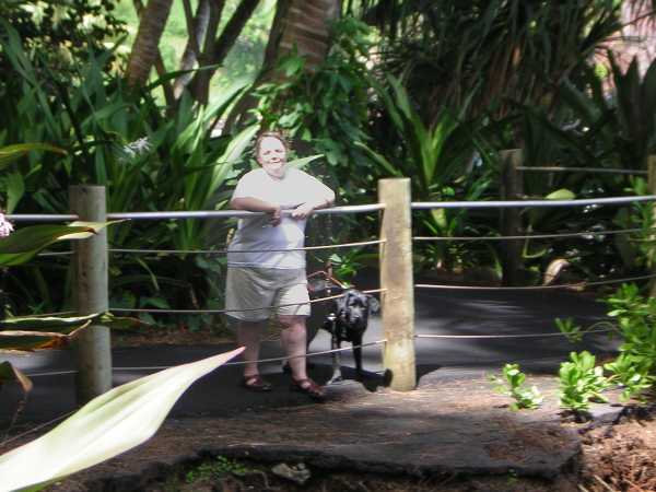 Maria in white t-shirt and tan shorts, her face a bit washed out by the sun, stands with forearms draped over a railing in front of a steep drop-off, with black lab Bella in harness at her side. Maria in white t-shirt and tan shorts, her face a bit washed out by the sun, stands with forearms draped over a railing in front of a steep drop-off, with black lab Bella in harness at her side.