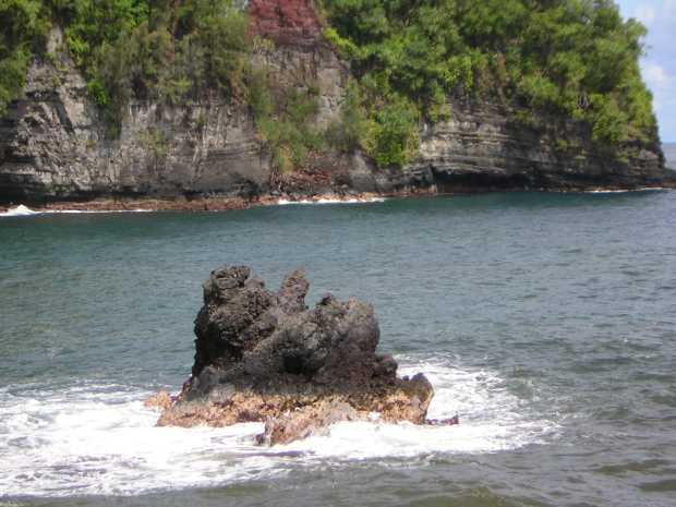 A large, craggy dark-grey rock, its base a larger tan, rises out of white sea foam and blue ocean water; beyond the water a massive grey and red rock cliff, obscured by hanging foliage, fills the view. A large, craggy dark-grey rock, its base a larger tan, rises out of white sea foam and blue ocean water; beyond the water a massive grey and red rock cliff, obscured by hanging foliage, fills the view.