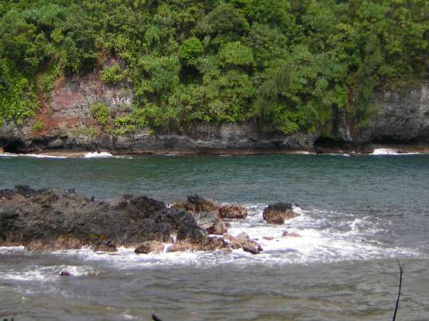 White foam and blue water lap against a low, sharp spur of black volcanic rock on three sides; beyond this stands a grey and red rock cliff, almost completely covered with green bushes. White foam and blue water lap against a low, sharp spur of black volcanic rock on three sides; beyond this stands a grey and red rock cliff, almost completely covered with green bushes.