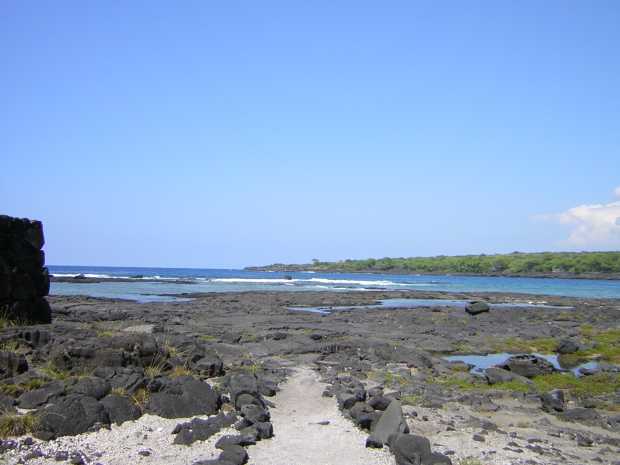 A white sandy path bordered by rough black cobbles leads away toward flat black lava and whitecapped surf beyond. The corner of a black stone wall is at left, and low forest extends out along the water at right. A white sandy path bordered by rough black cobbles leads away toward flat black lava and whitecapped surf beyond. The corner of a black stone wall is at left, and low forest extends out along the water at right.
