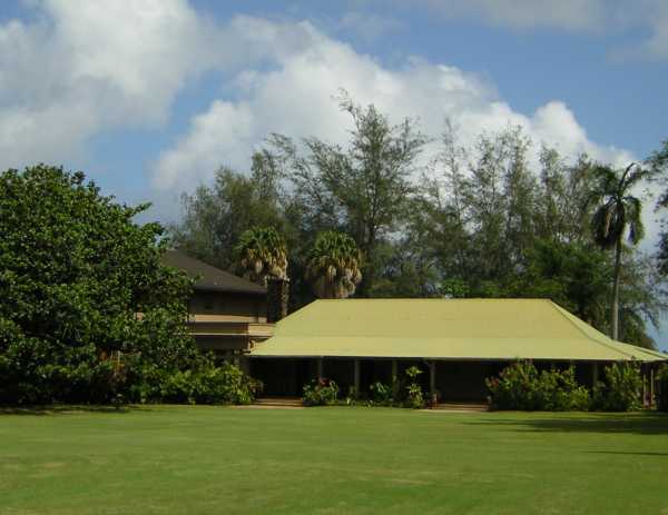 Beyond a vast expanse of lawn, a broad, low green roof topping a dark structure extends outward from the left of a higher, more grand building that is mostly obscured by a huge thick-leafed monkeypod tree. Beyond a vast expanse of lawn, a broad, low green roof topping a dark structure extends outward from the left of a higher, more grand building that is mostly obscured by a huge thick-leafed monkeypod tree.