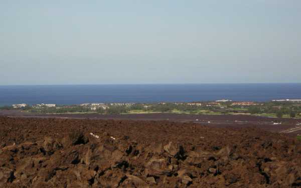 Looking down from a height, a reddish-brown jumble of rough rocks gives way to darker rock, then to a distant green swath of vegetation dotted with white and red buildings; beyond the sea stretches to the horizon. Looking down from a height, a reddish-brown jumble of rough rocks gives way to darker rock, then to a distant green swath of vegetation dotted with white and red buildings; beyond the sea stretches to the horizon.