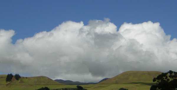 Velvety olive-green hills rise abruptly from a green plain against a backdrop of fluffy grey-white clouds topped with deep blue sky. Velvety olive-green hills rise abruptly from a green plain against a backdrop of fluffy grey-white clouds topped with deep blue sky.