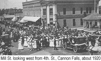 Black and white  photograph of Mill St. looking west from 4th. St., Cannon Falls, about 1920, showing buildings presumably owned by Dick Dibble, with a crowd of people celebrating a patriotic holiday